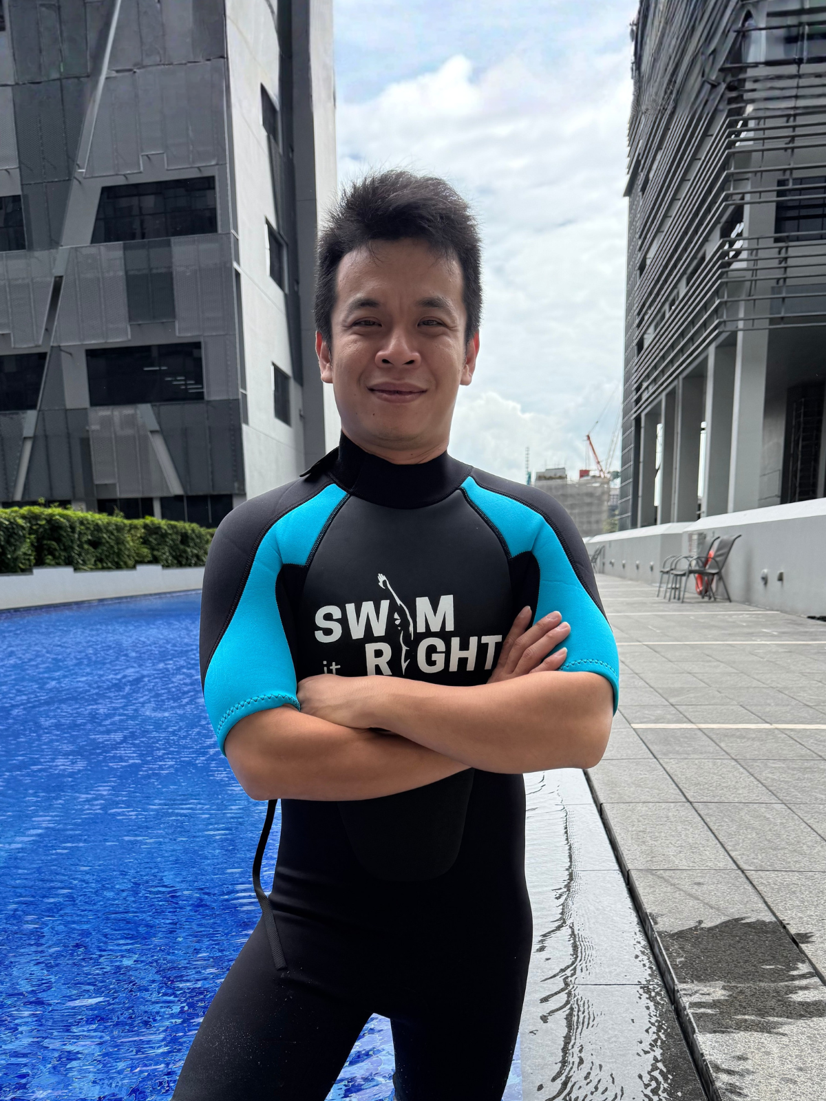 Man standing in a swimming pool with crossed arms, wearing a black and blue wetsuit, on an urban rooftop with modern buildings background.
