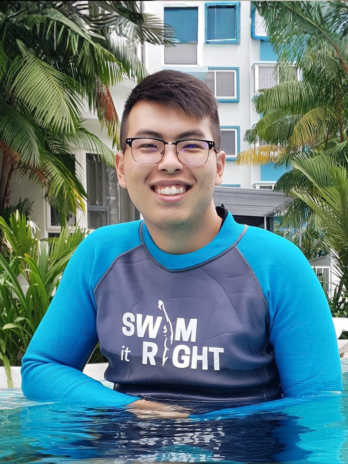 A young man sitting on the edge of a swimming pool with his feet in the water, wearing a swim shirt, shorts, glasses, and a cap, surrounded by greenery and trees.