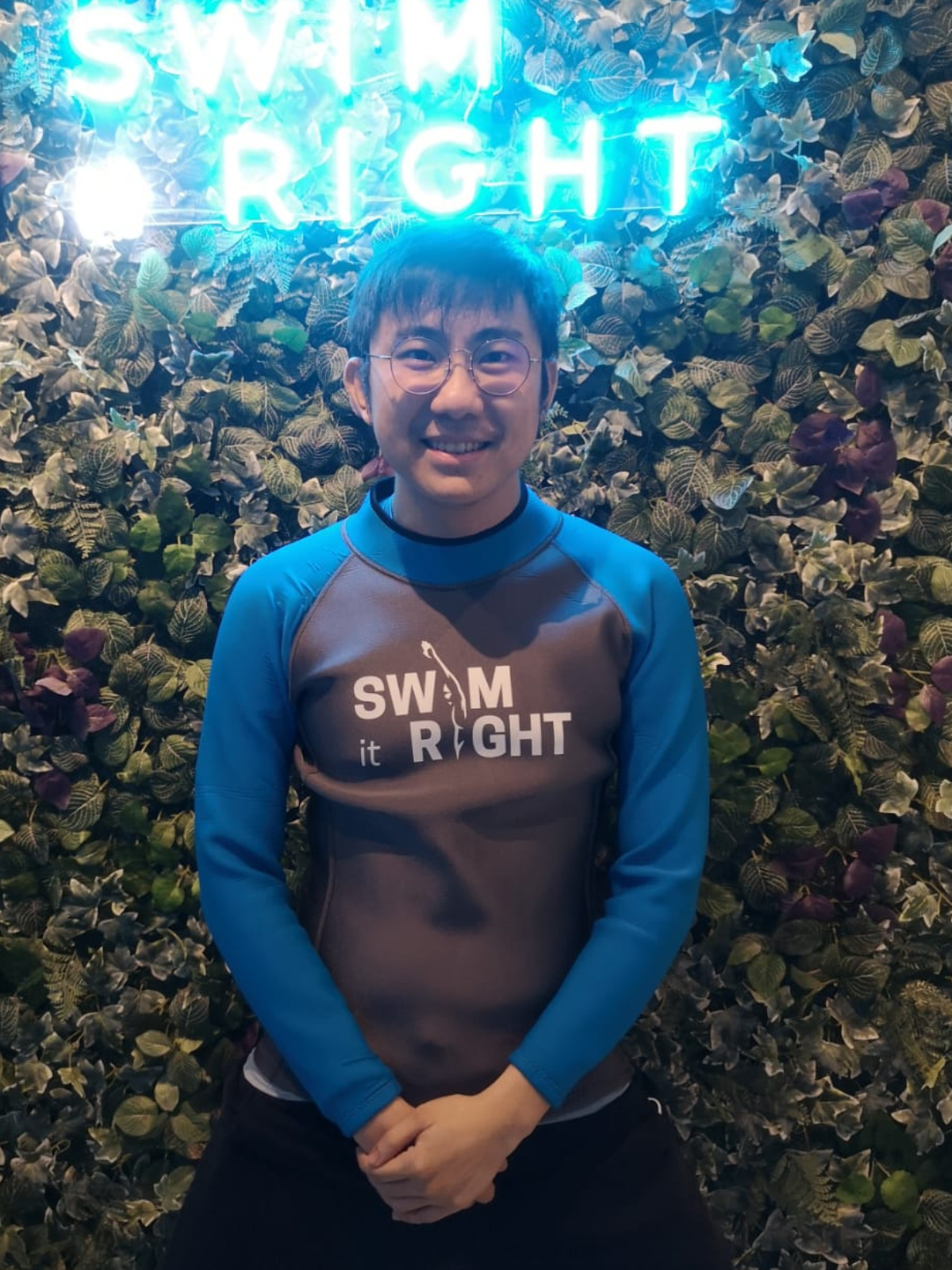 Smiling young man wearing glasses and a swim-themed athletic shirt, standing in front of a leafy background with blue neon lighting.