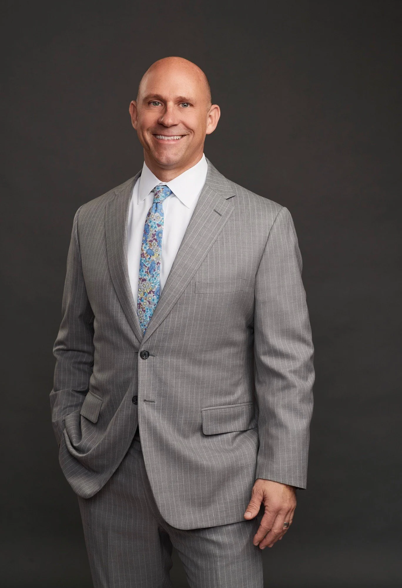 Man in a light gray suit with a patterned tie smiling and standing against a dark background.