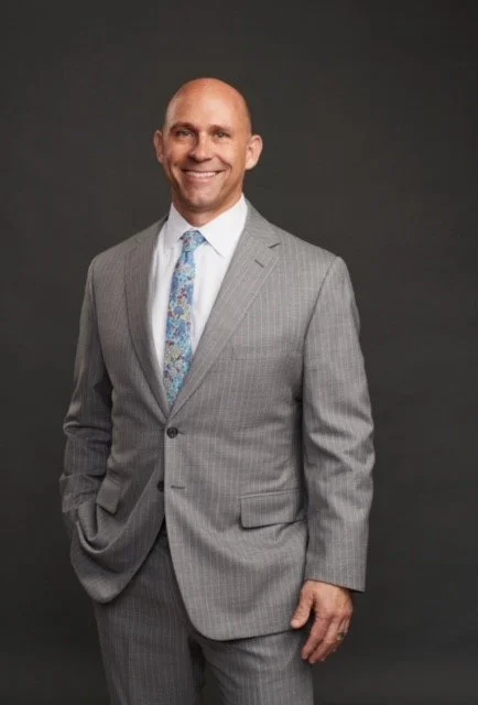Man in a light gray suit with a patterned tie smiling and standing against a dark background.
