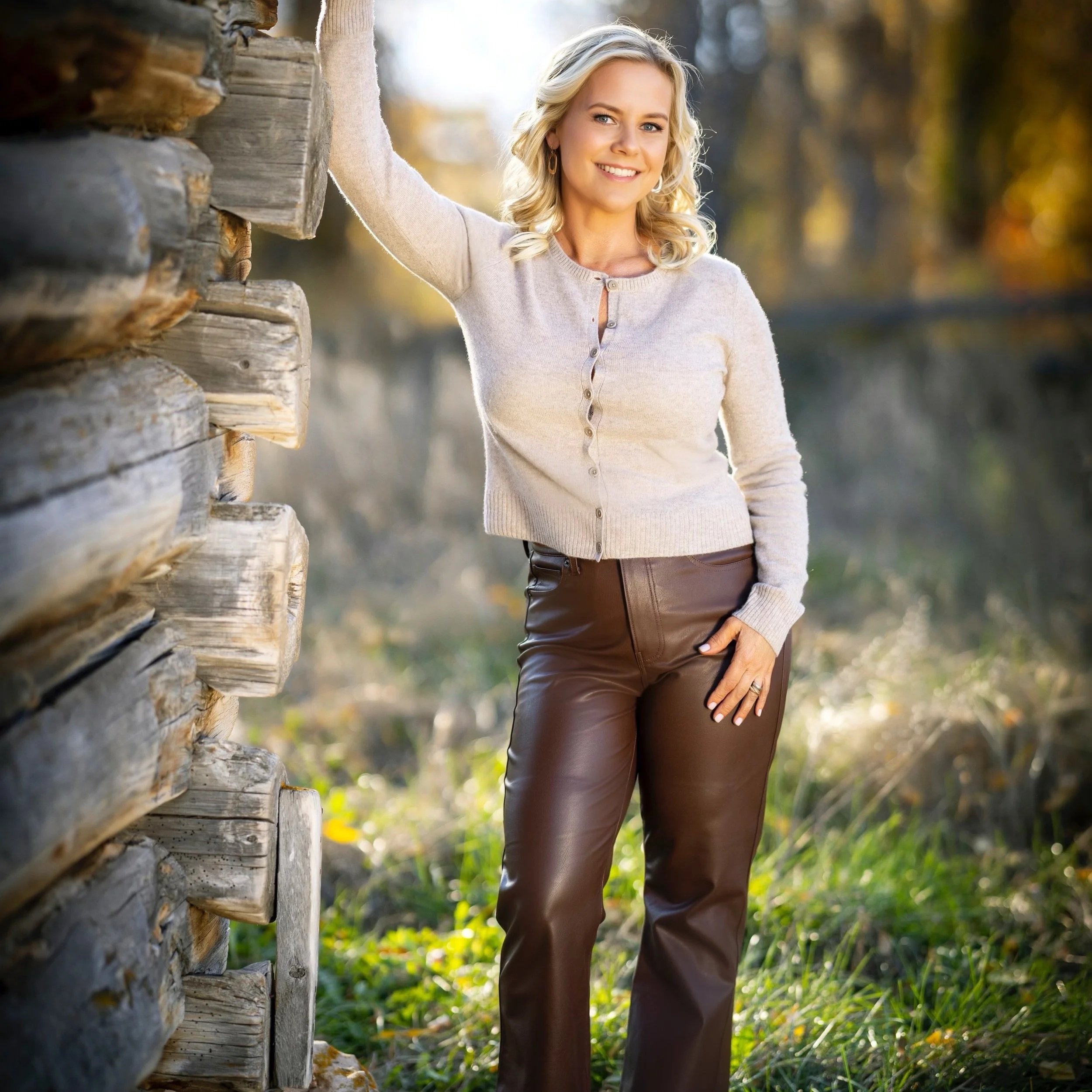 Patricia Kennedy, founder of Rendezvous Design, standing outdoors beside a weathered log cabin wall
