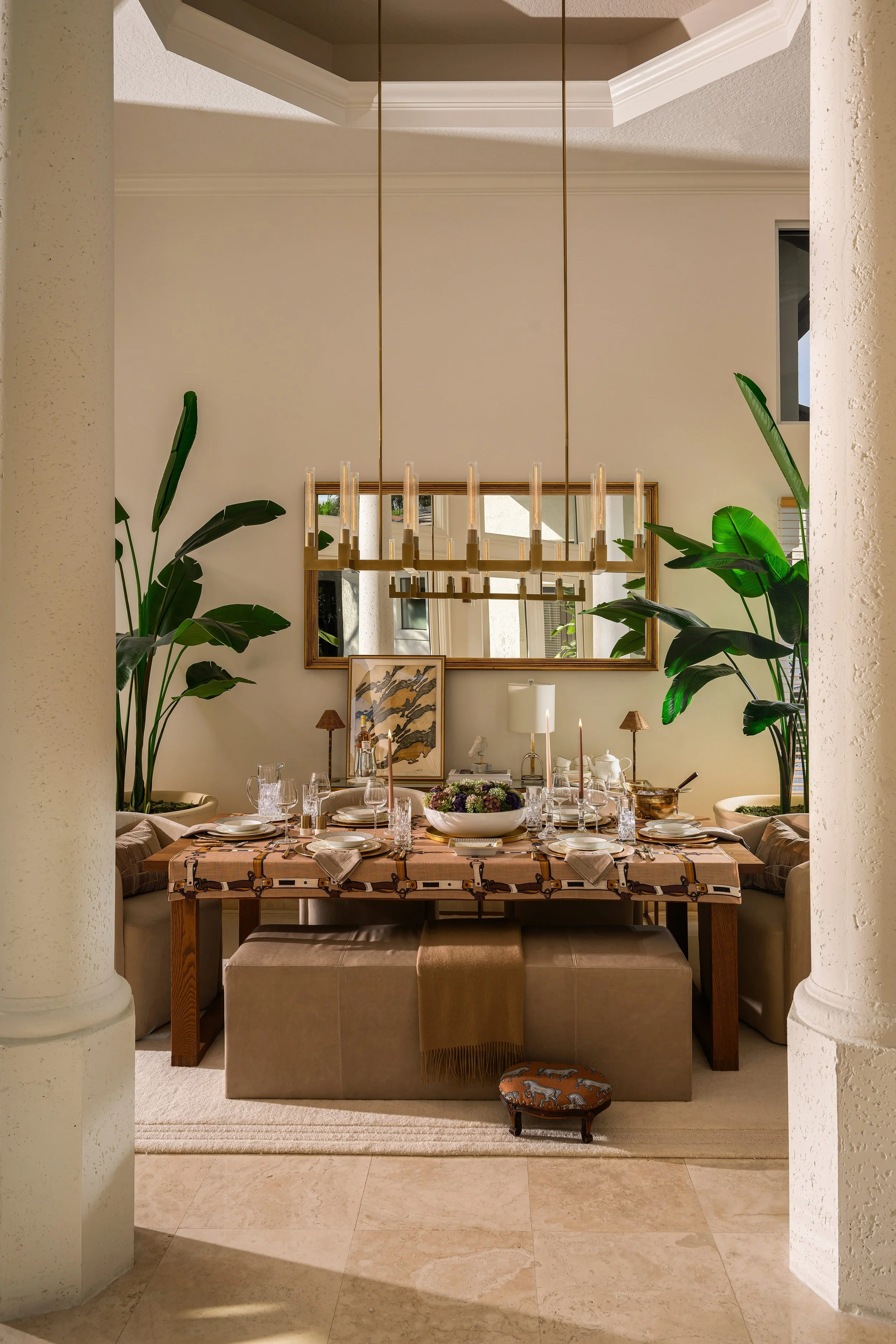 Dressage Dining room framed by columns: wood table with equestrian strap runner, upholstered benches, brass chandelier, and tropical plants.