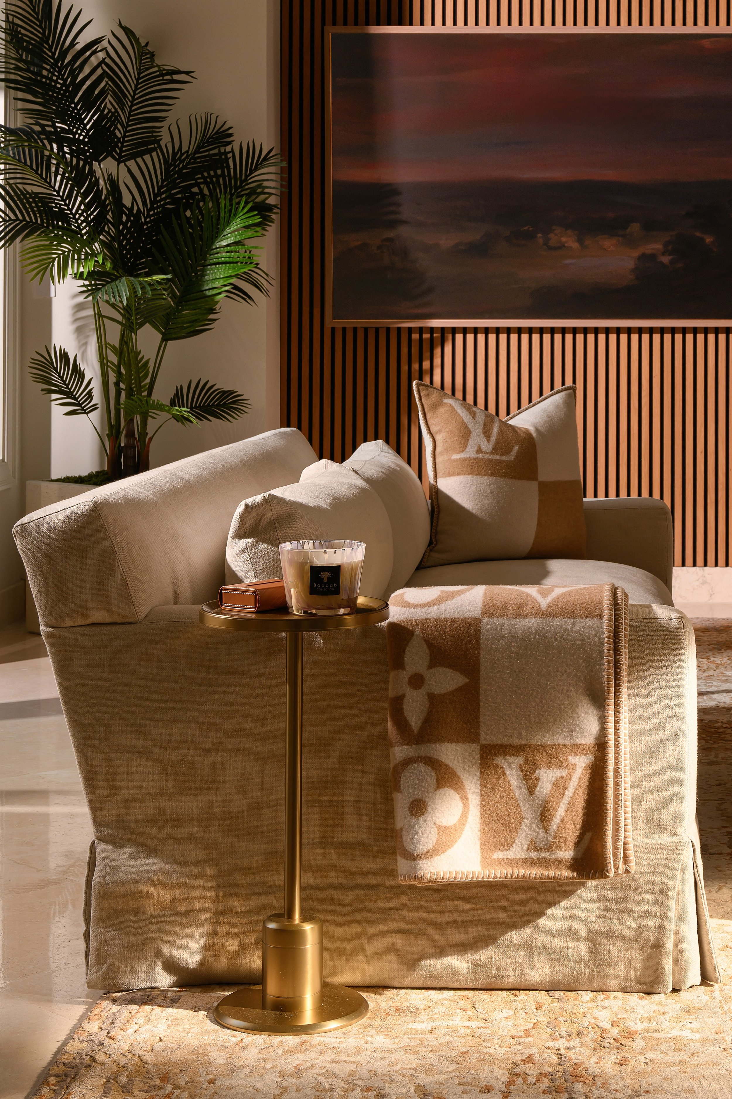 Modern Marvel vignette: linen slipcovered chair with Louis Vuitton throw and monogram pillow, brass candle table, palm, and landscape painting.
