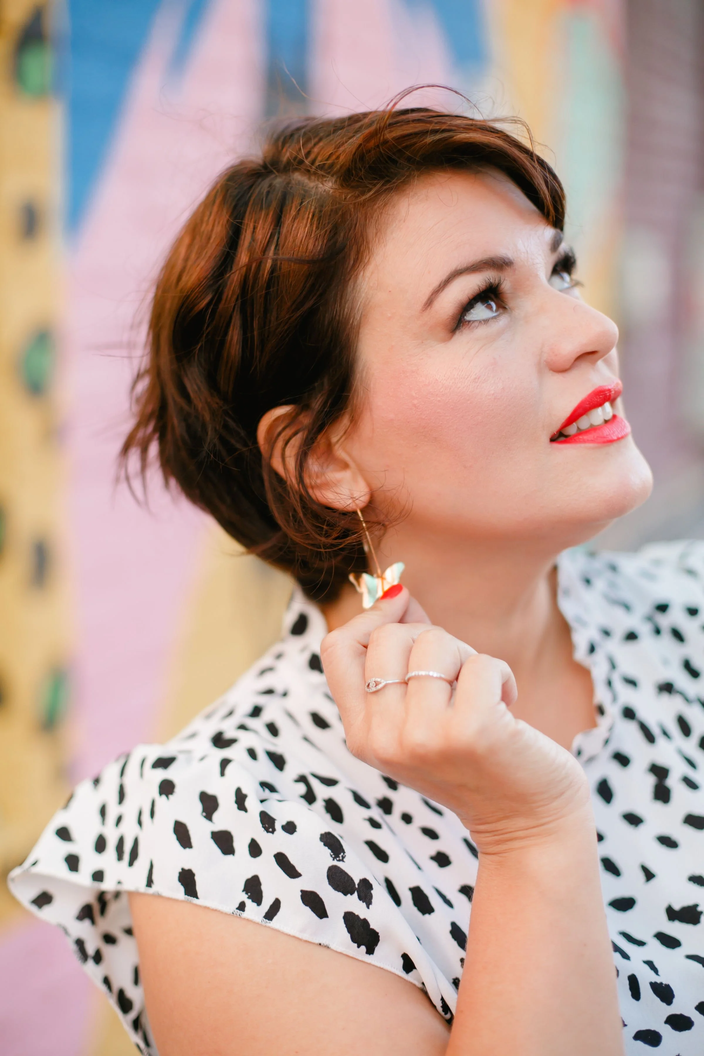 Woman with short wavy brown hair and red lipstick looking upward, wearing a polka dot top and butterfly-shaped earrings