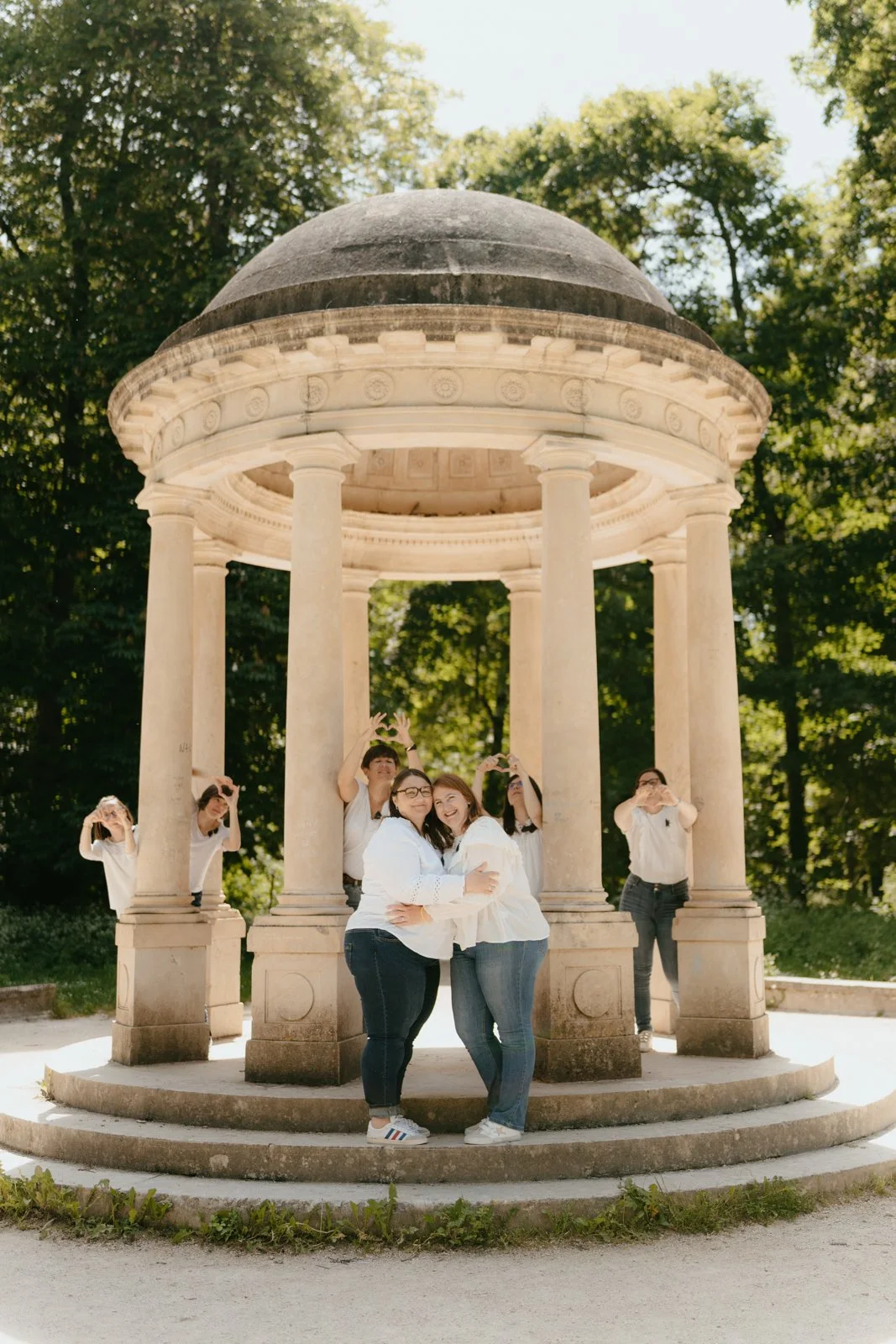 Groupe de sept jeunes souriants posant devant un petit pavillon rond en pierre avec des colonnes, entouré d'arbres verts en été.