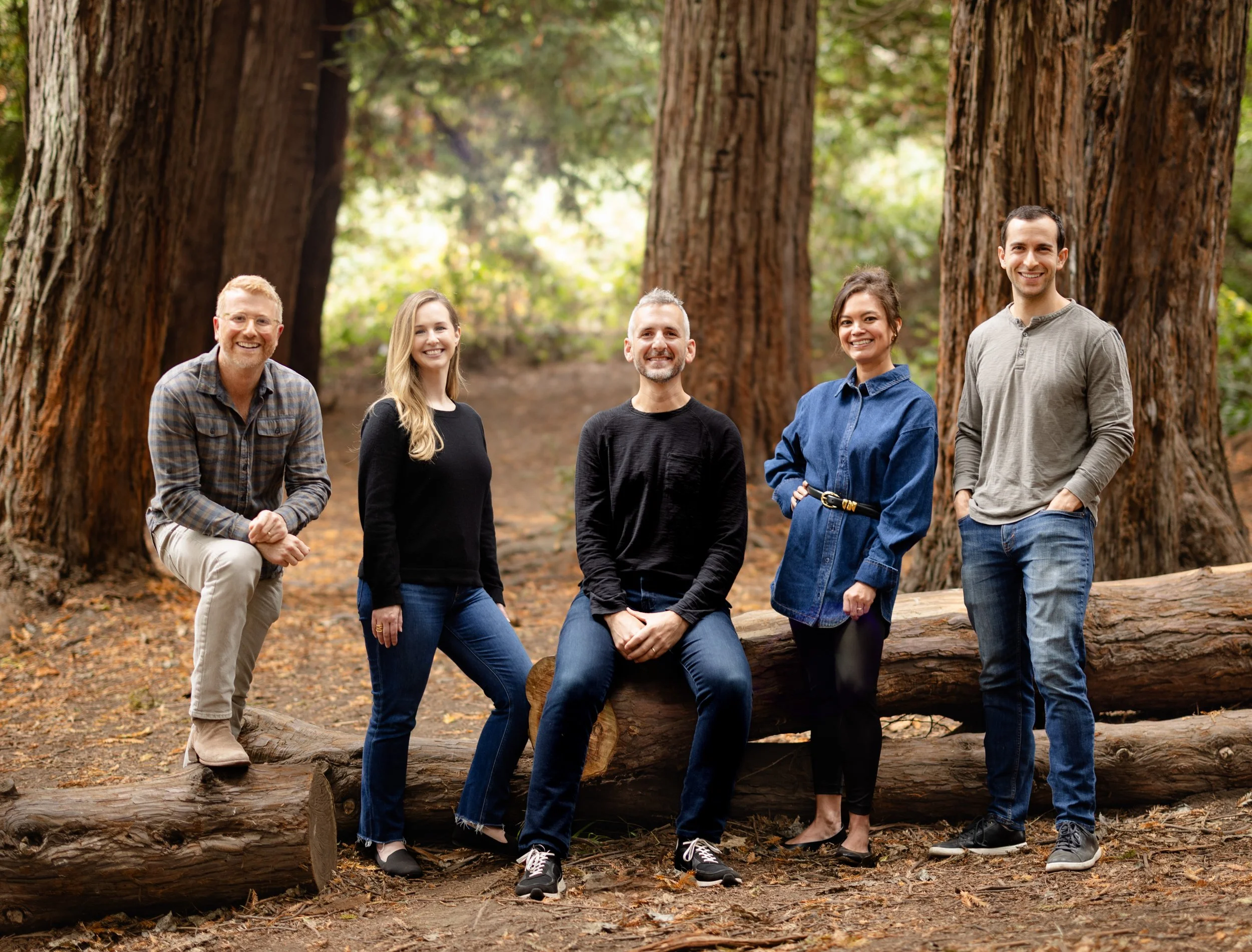 A group of five people stands and sits on logs in a forest, smiling warmly.