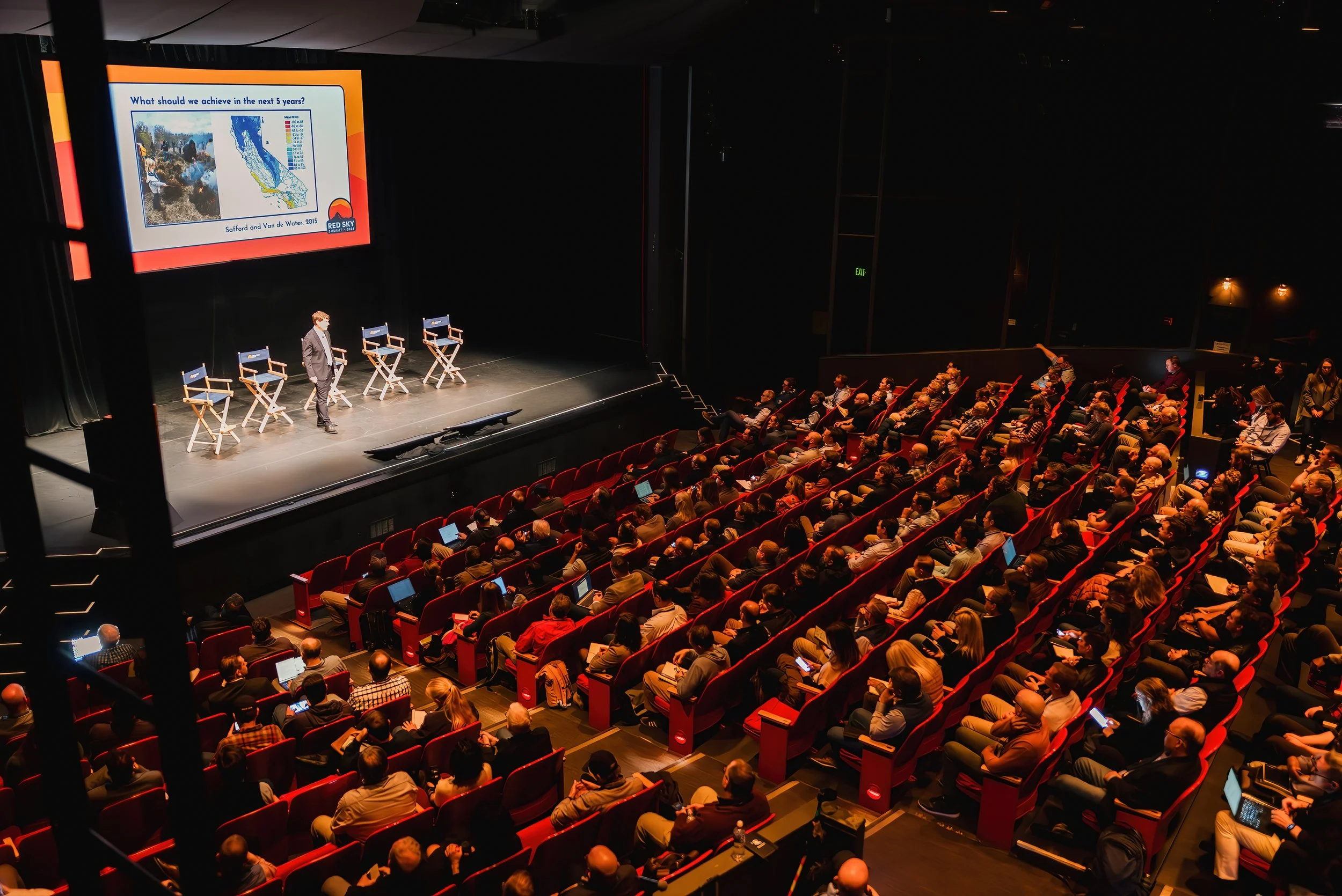 A presenter on stage addresses a packed auditorium, next to a large screen displaying a slide.
