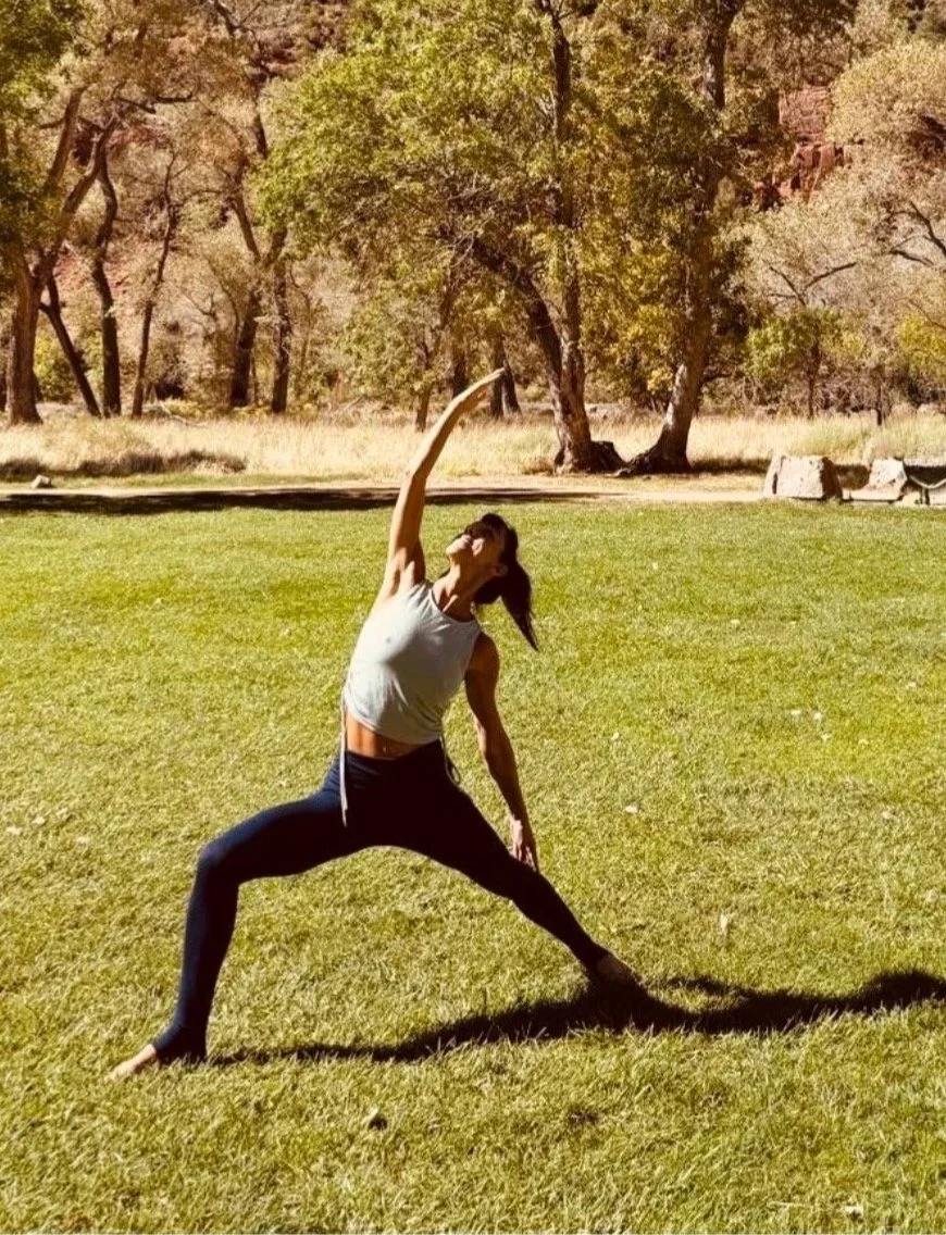 A person practicing yoga outdoors on a grassy field, surrounded by trees with autumn foliage, twisting in a yoga pose.