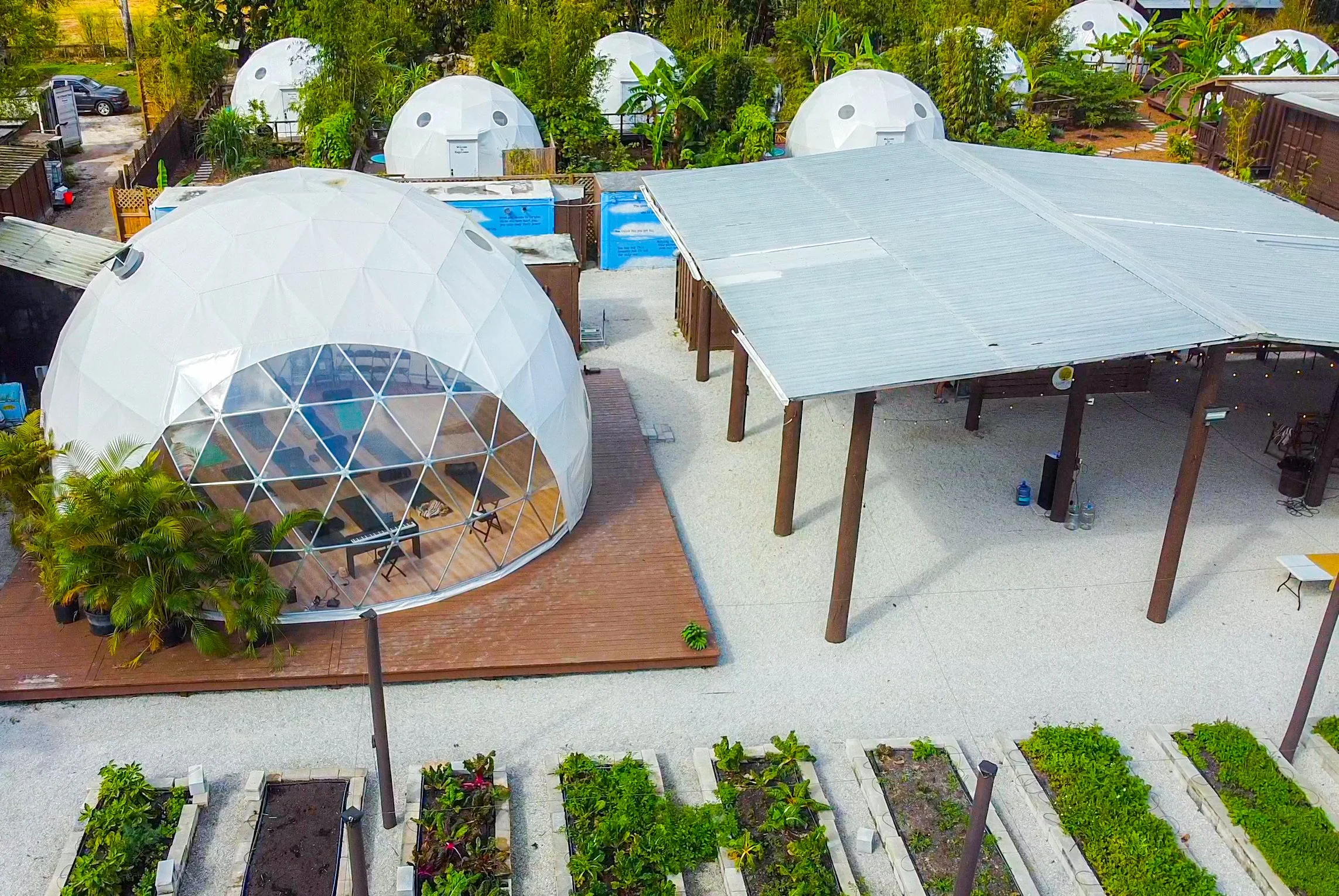 Aerial view of a hostel or retreat with geodesic dome structures, a covered open space, and garden beds.