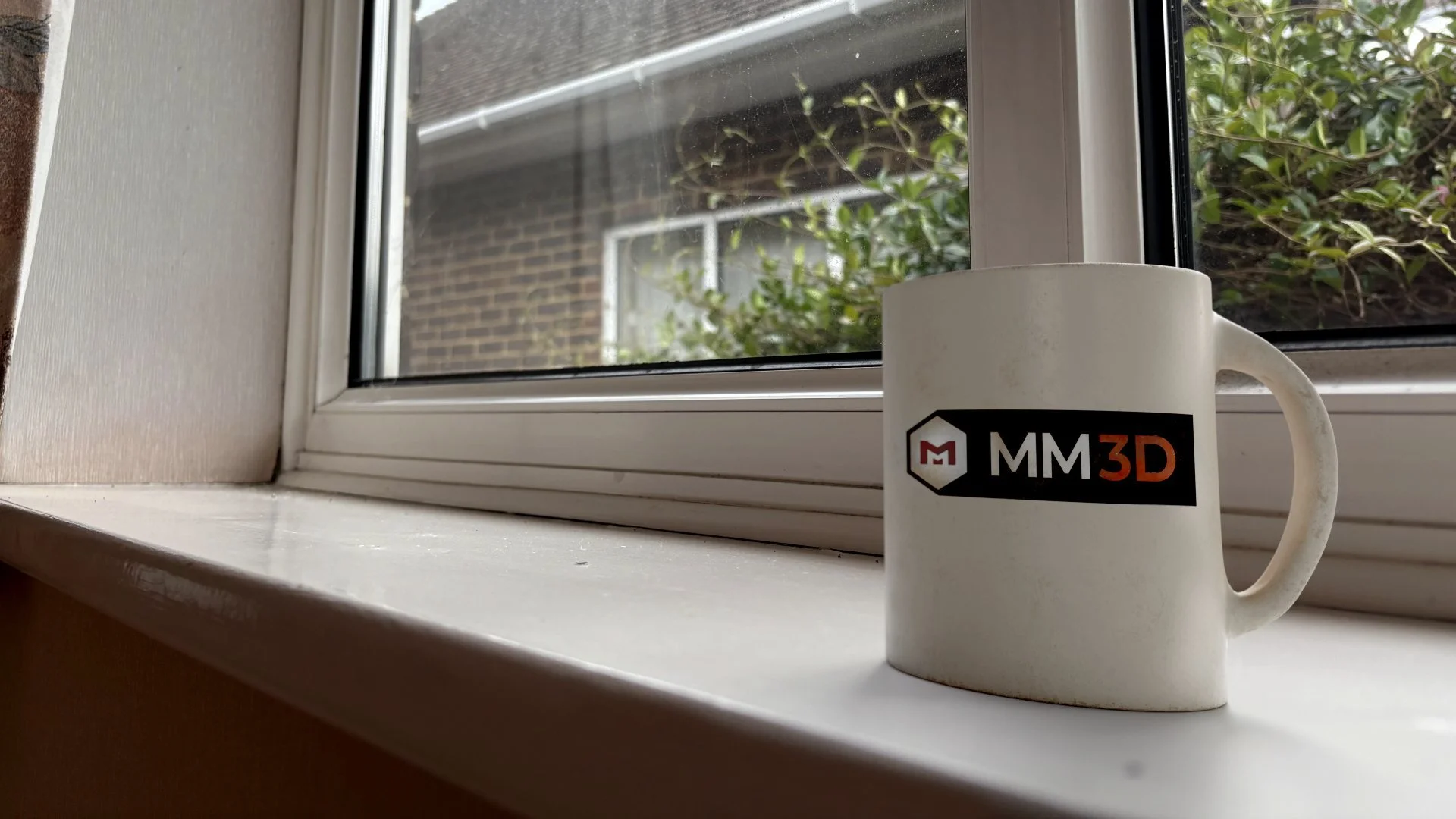 A white coffee mug with a logo that says 'MM3D' in black and orange, placed on a windowsill with a view of a brick building and greenery outside.