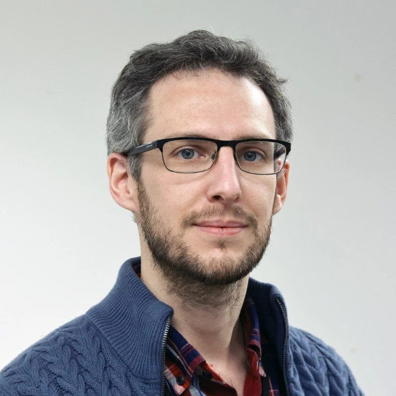 A man with glasses, a beard, and short dark hair, wearing a blue zip-up sweater over a plaid shirt, standing against a plain light background.
