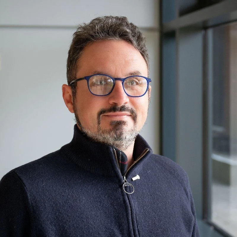 A man with glasses and a beard wearing a dark zip-up sweater standing indoors near a window.