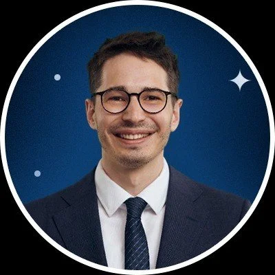 A young man with glasses and a suit smiling against a blue backdrop with stars.