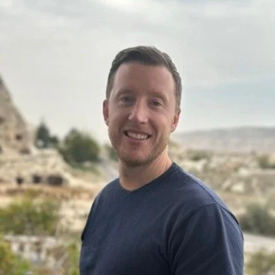 A smiling man with short dark hair, wearing a navy blue shirt, standing outdoors with a blurred rocky landscape and cloudy sky in the background.