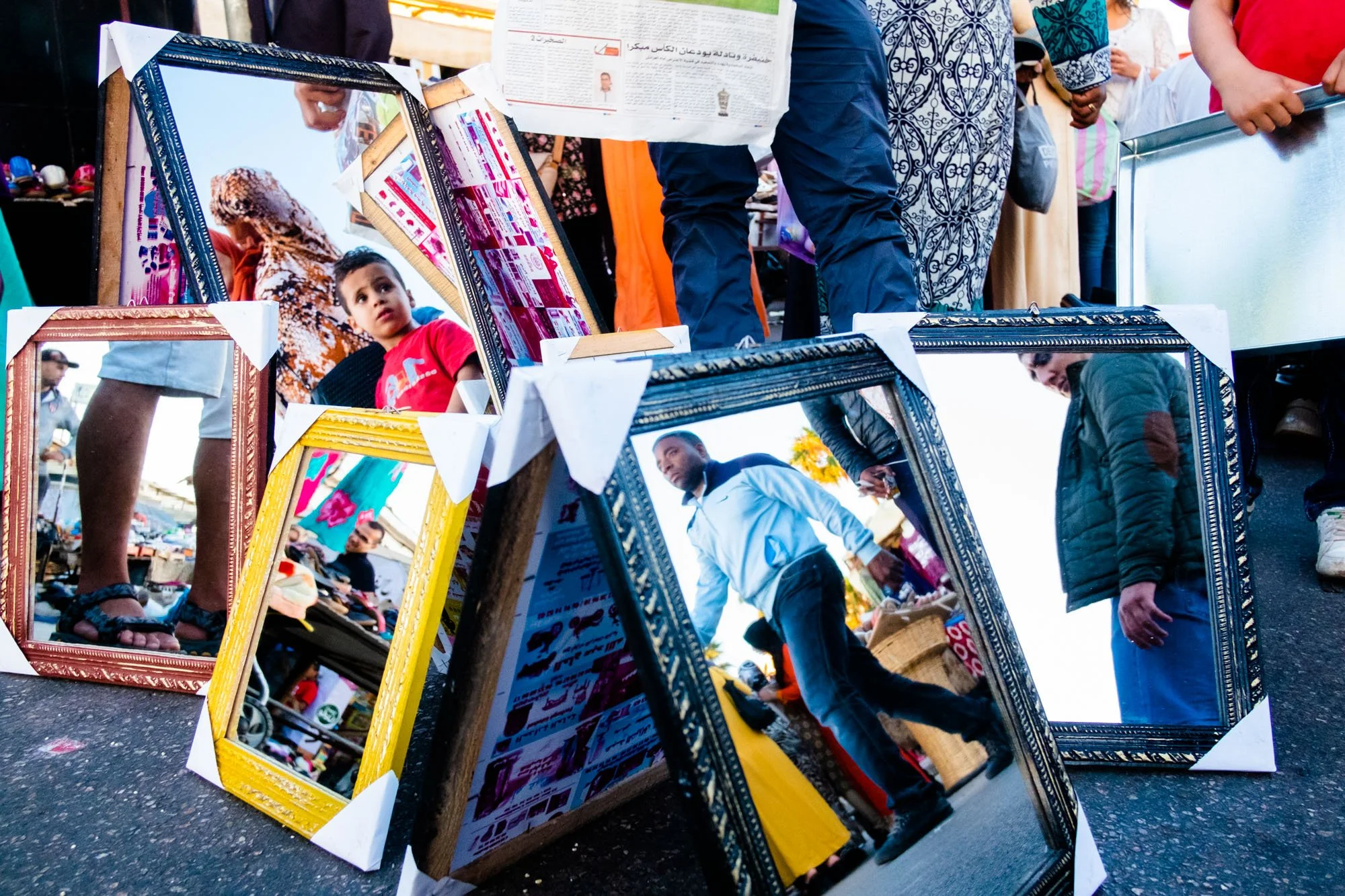 A dozen second-hand mirrors and picture frames propped on flea-market tarmac at odd angles, each catching a different slice of the market above — sandalled feet, a man in a pale blue jacket mid-stride, a woman in a patterned djellaba, an Arabic newspaper; only legs and hands of the real crowd appear above the frames