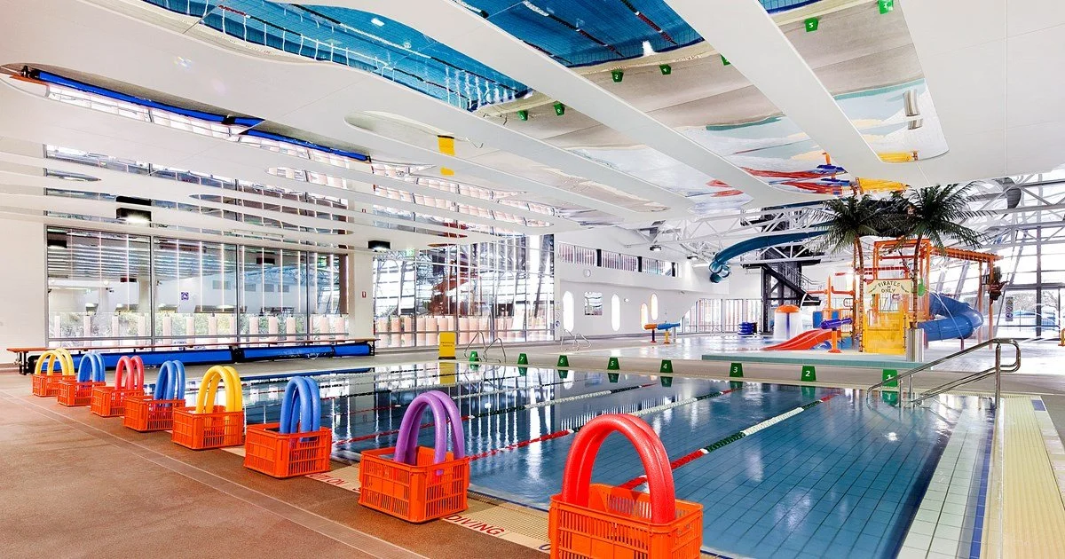 Indoor swimming pool with colorful floatation rings stored in orange baskets by the poolside, water slides, palm trees, and a modern, bright architectural design with large windows.