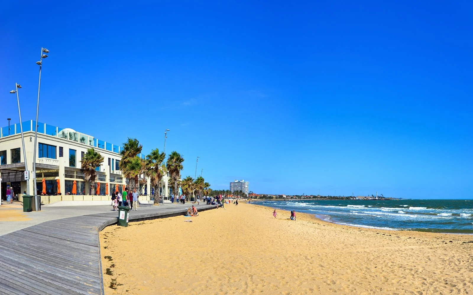 Beach with sandy shoreline, blue ocean waves, palm trees along a promenade, people walking and sitting, and buildings in the distance under a clear blue sky.