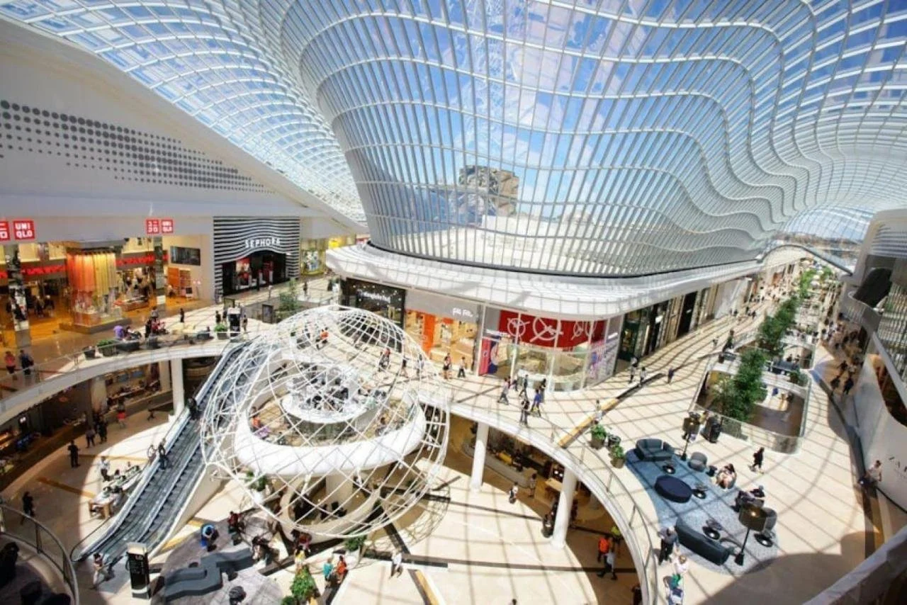 Interior view of a modern shopping mall with multiple levels, a glass dome structure, escalators, and various retail stores and shoppers.