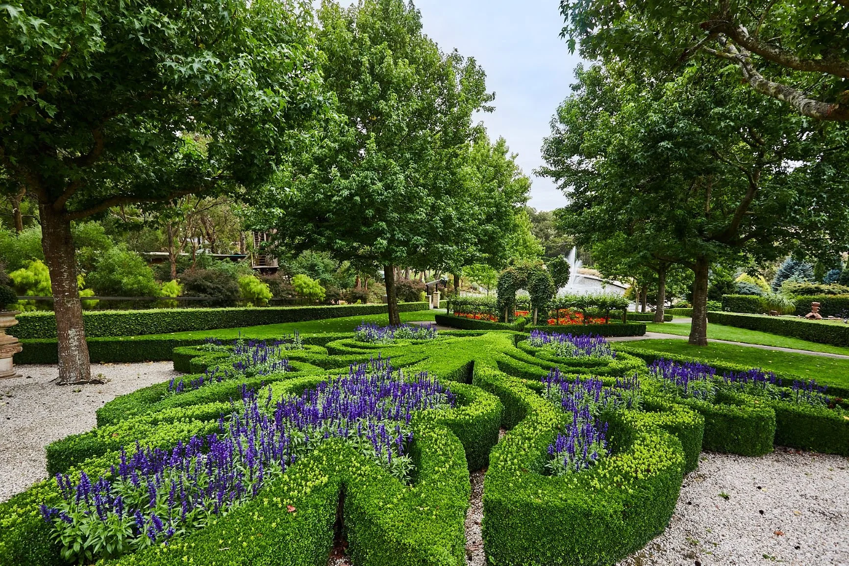 Well-maintained garden with neatly trimmed hedges, purple flowers, tall green trees, and a fountain in the background.