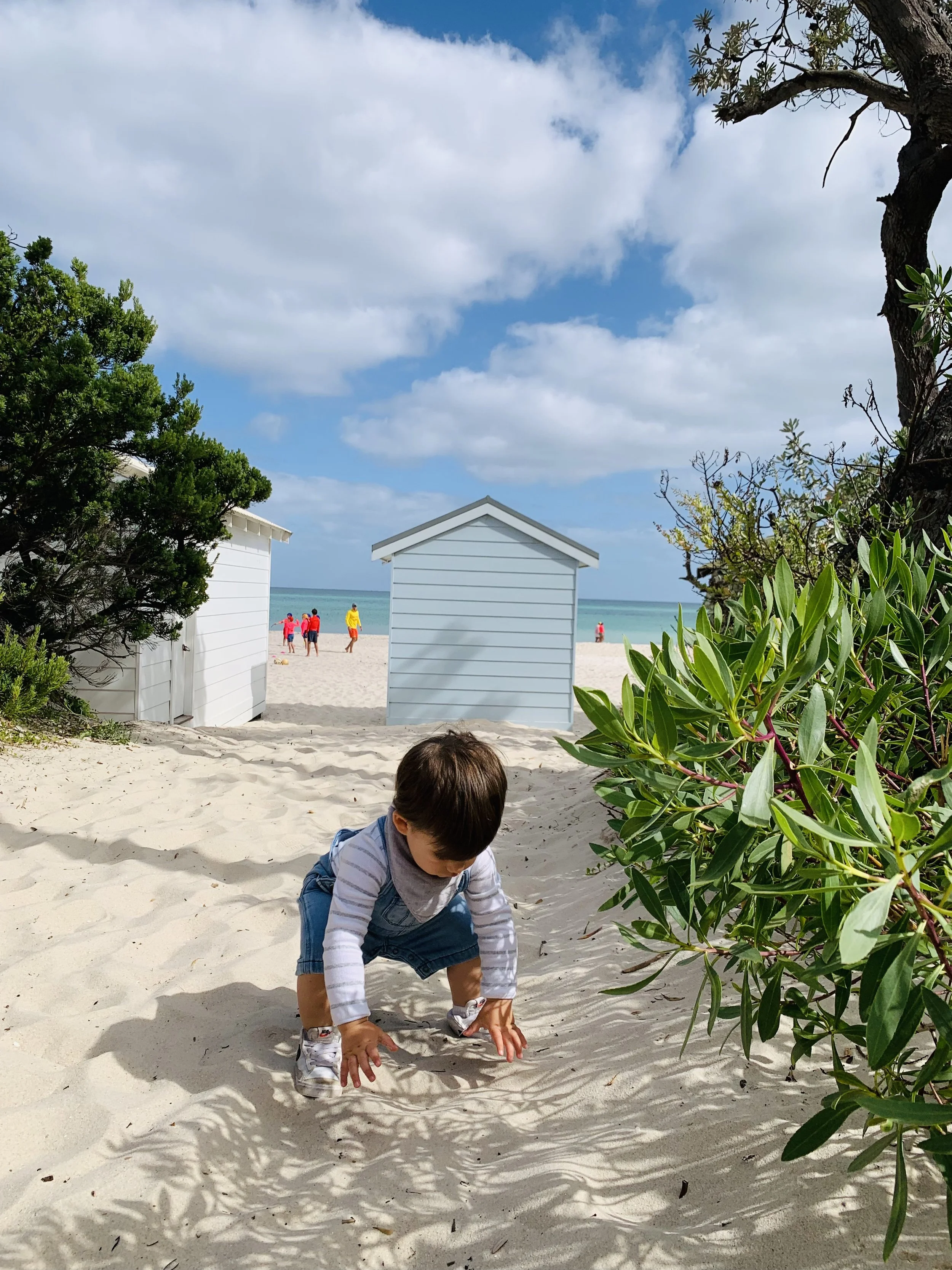 A young boy crawling on sandy beach with two small white beach houses and green bushes in the background, blue sky with clouds, and people by the ocean.