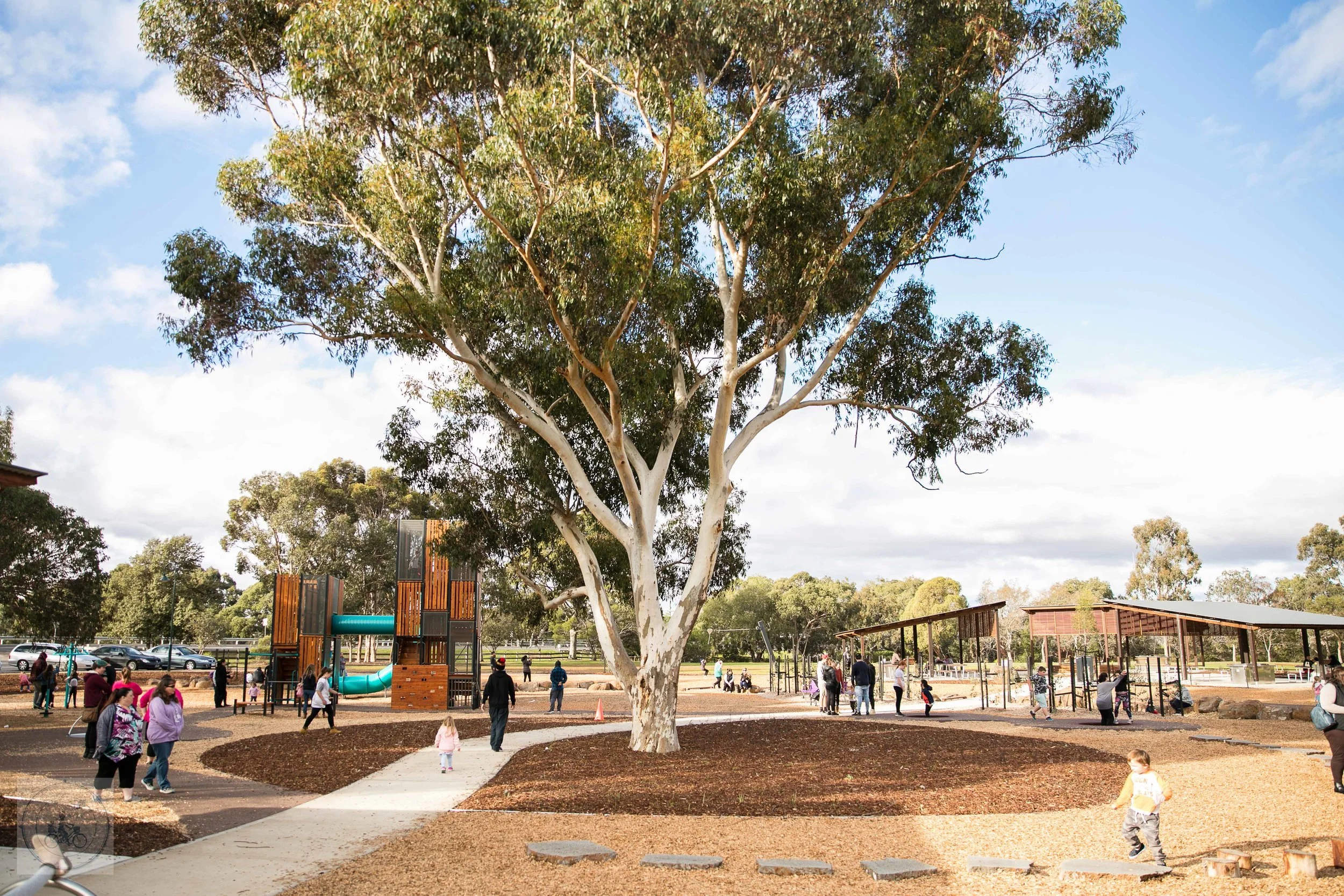 Public park with children and adults, large tree in center, playground structures, covered seating area, open sky, and trees in the background.