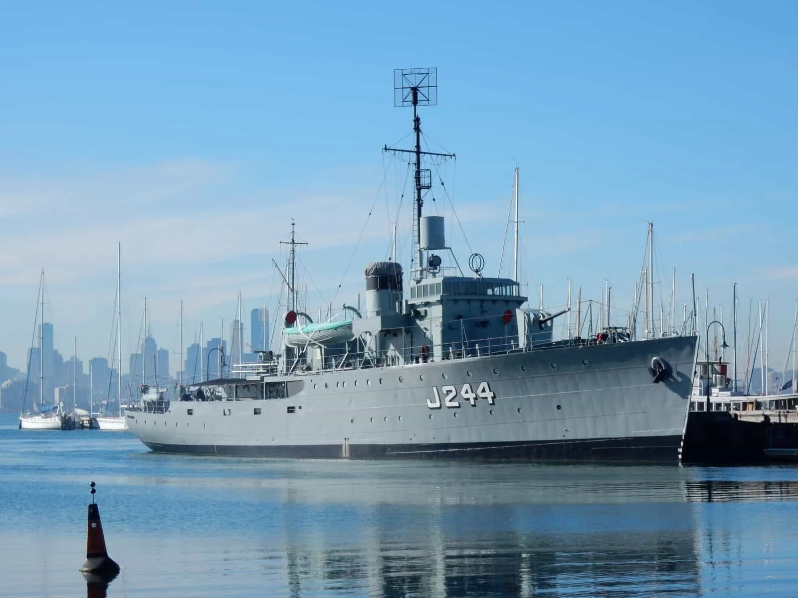 A military ship docked at a marina with a city skyline in the background, under a clear blue sky.