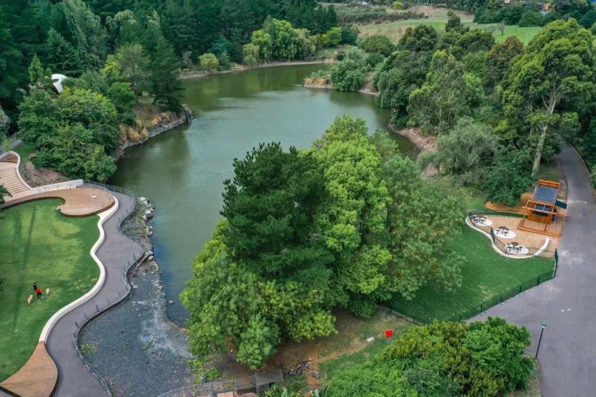 Aerial view of a park with a pond, surrounded by green trees and landscaped areas, including pathways, a small outdoor seating area, and a man walking his dog.