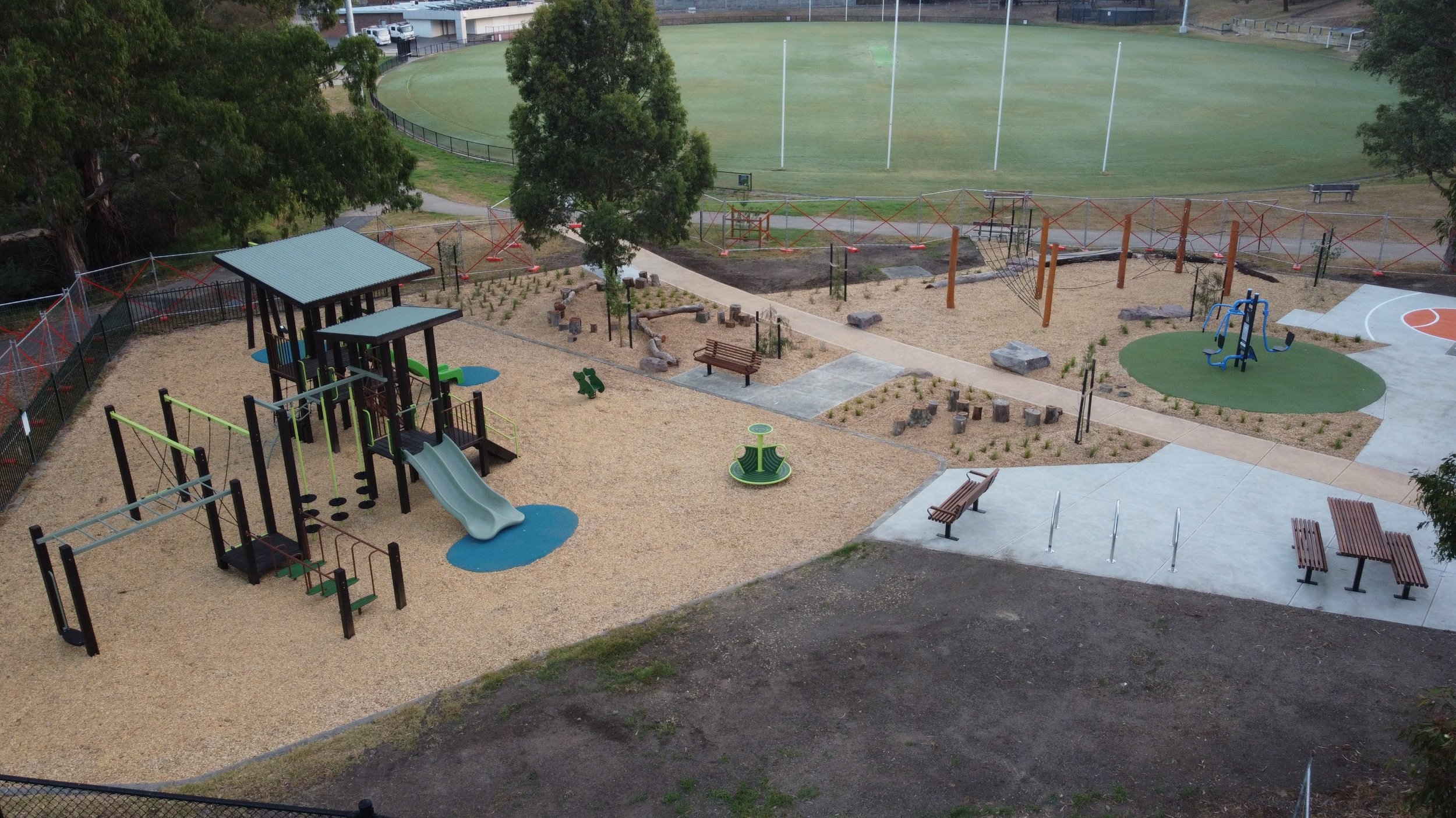 Children's playground with slides, swings, and climbing structures, surrounded by trees, benches, and walking paths, with a sports field in the background.