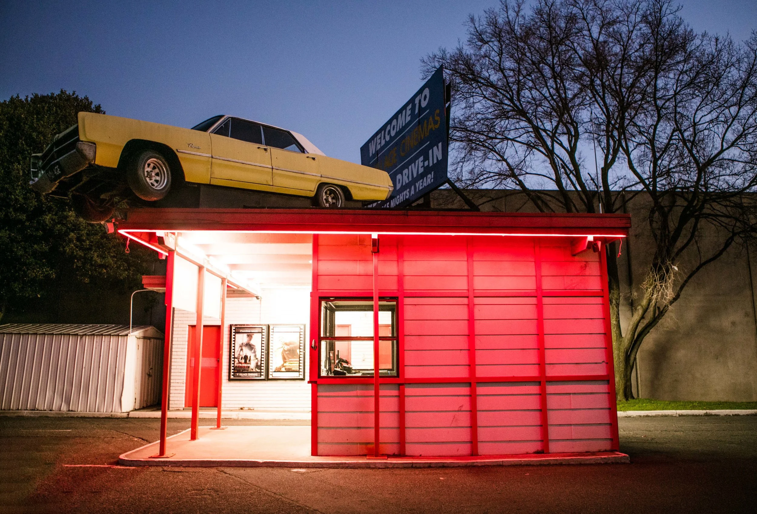 A small fast food or concession stand building painted in red with neon lighting at night. On the roof, there is a yellow vintage car mounted as a decorative sign. Behind, there is a billboard with partially visible text and a silhouette of leafless trees.
