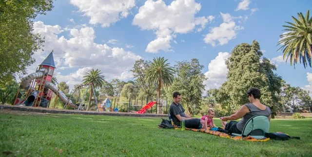 Family having a picnic on the grass at a park with a playground and palm trees in the background.