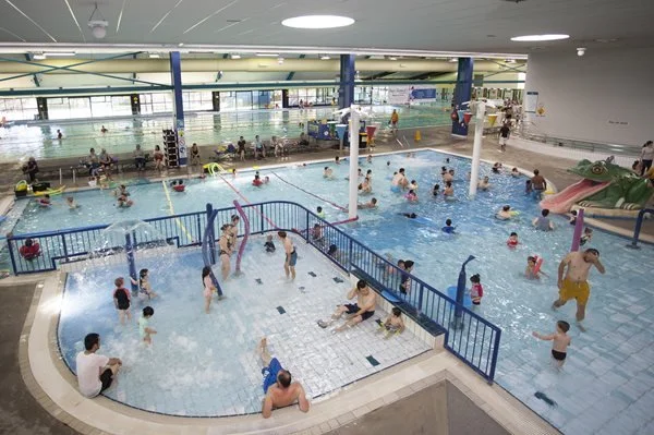Indoor swimming pool filled with children and adults swimming, playing, and relaxing. There are slides, water features, and a designated shallow area with a blue safety fence.