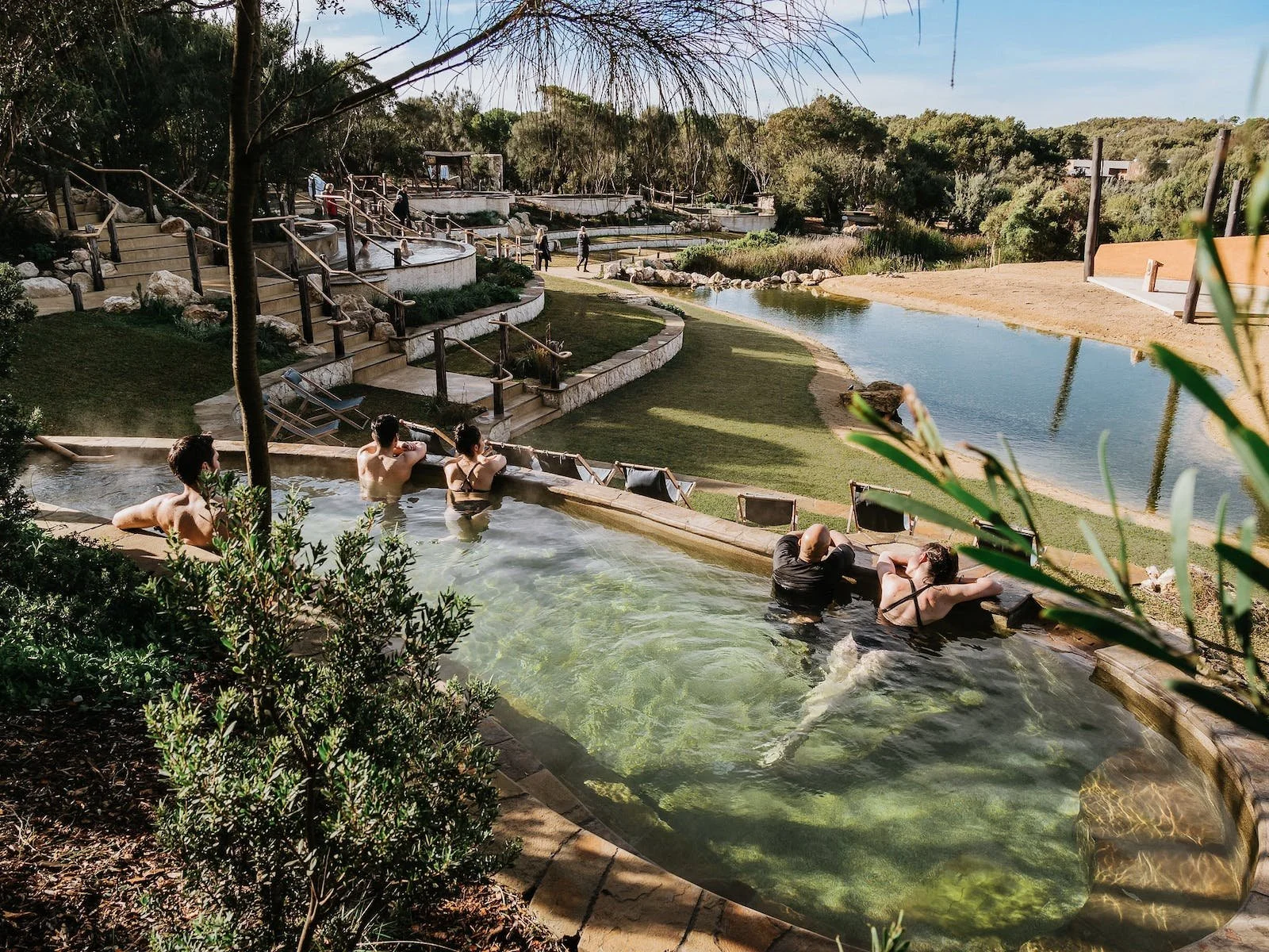 People relaxing in hot spring pools surrounded by greenery, rocks, and a scenic landscape with a body of water and trees in the background.