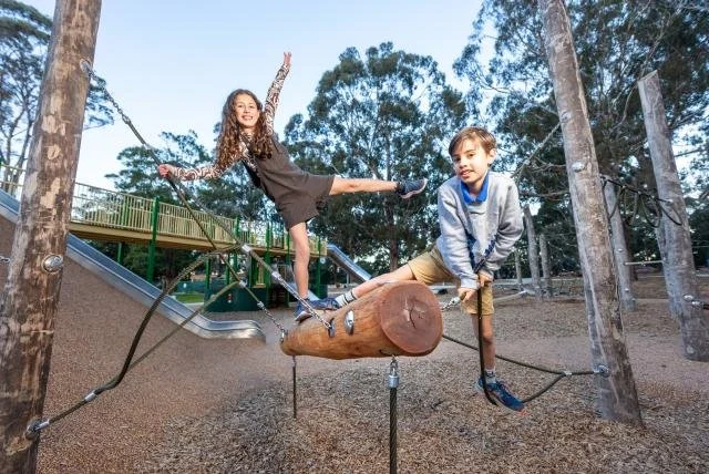 A girl and a boy playing on a balance beam at a playground, surrounded by trees.