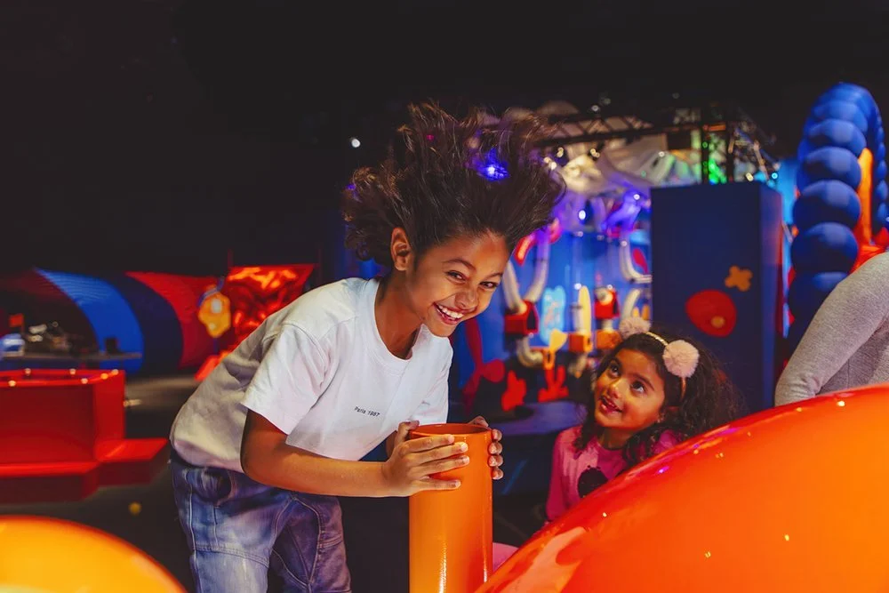Two girls with curly hair, one older and one younger, playing and smiling at an indoor arcade or entertainment center with colorful lights and structures.