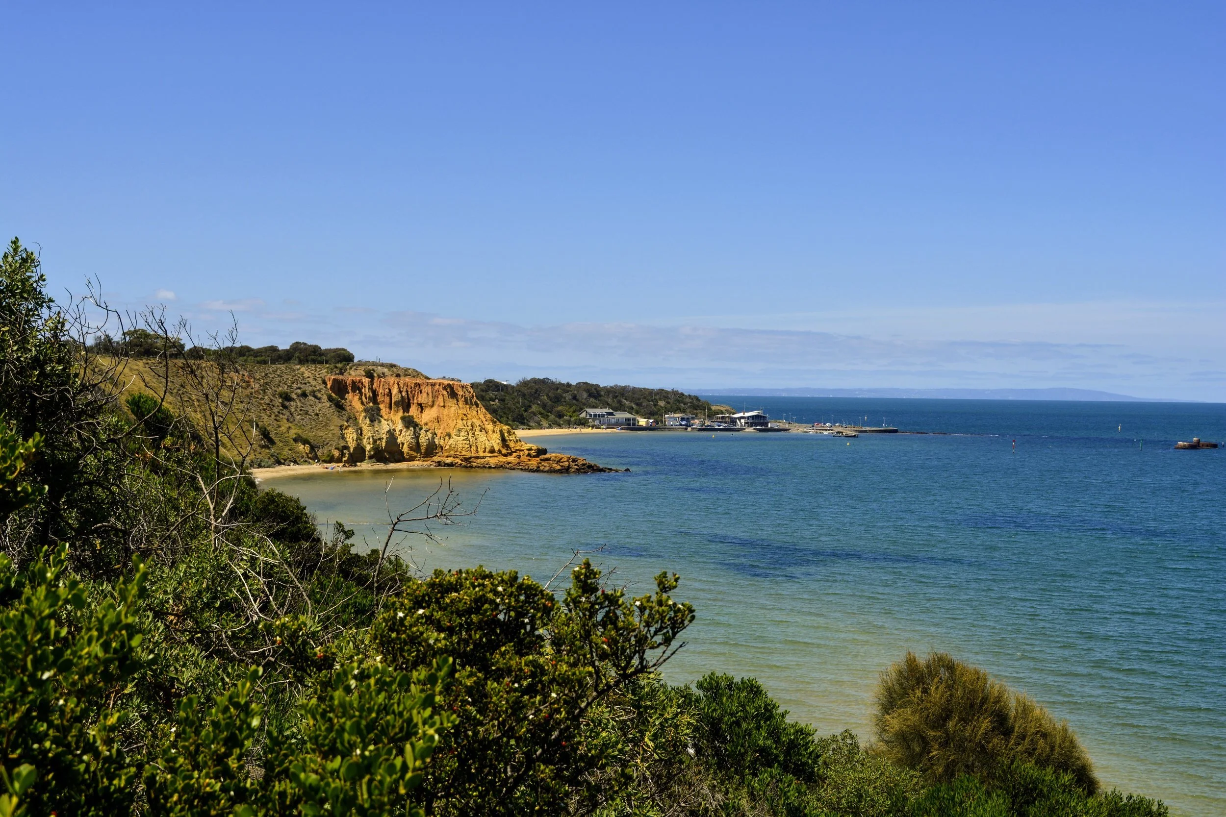 Coastal landscape with sandy beach, rocky cliffs, greenery, and calm blue ocean under a clear sky.