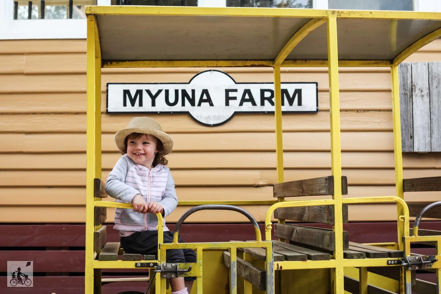 A young girl sitting on a yellow wooden cart in front of a sign that says Myuna Farm. She is wearing a straw hat and smiling.