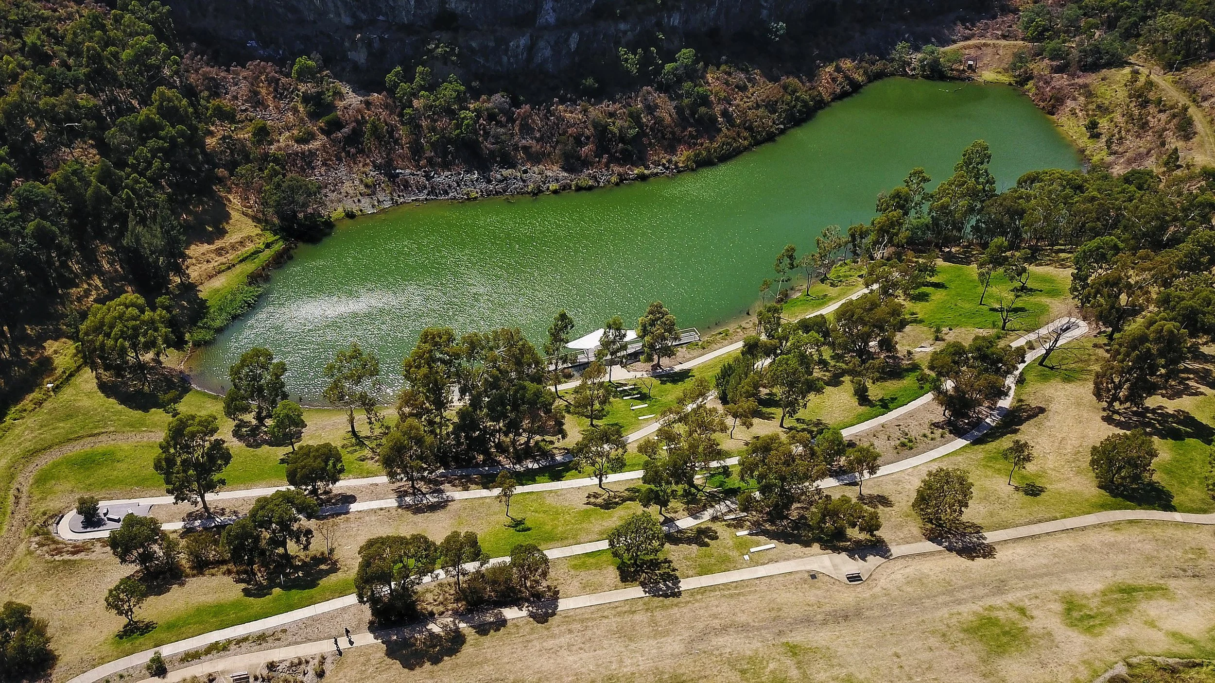 Aerial view of a park with winding pathways, scattered trees, and a large green lake surrounded by rocky hills.