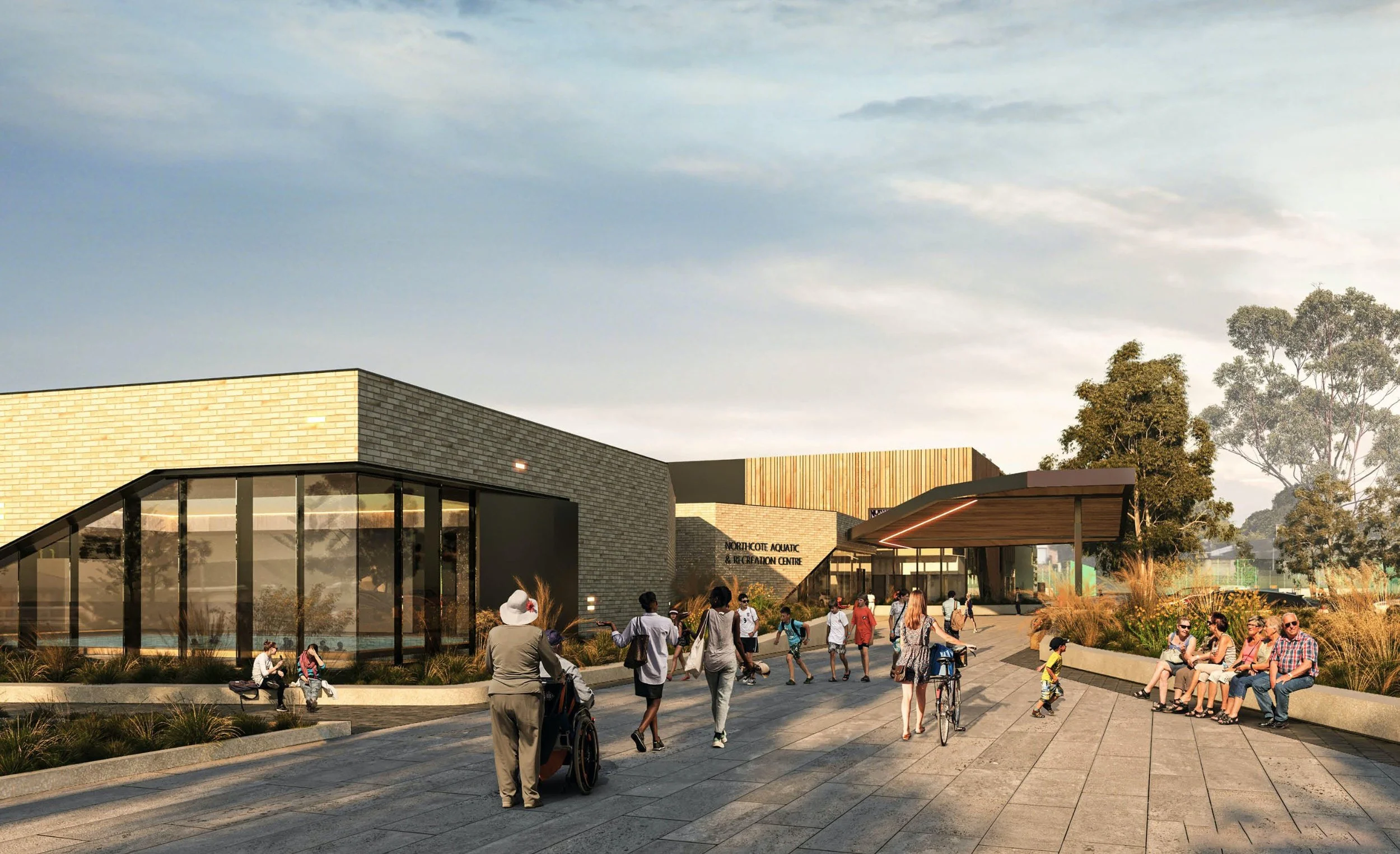 People gathered outside the Northcote Aquatic & Recreation Centre, with some sitting on benches, some walking, and others riding bikes, under a modern architectural building with large glass windows and a wooden canopy, surrounded by landscaped plants and trees.