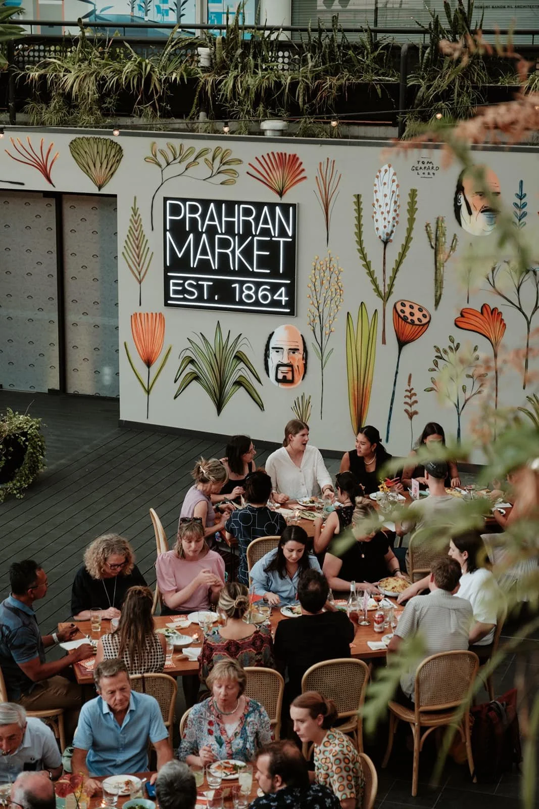 People dining at tables inside Prahran Market, established in 1864, with a decorative wall featuring illustrated plants and faces in the background.