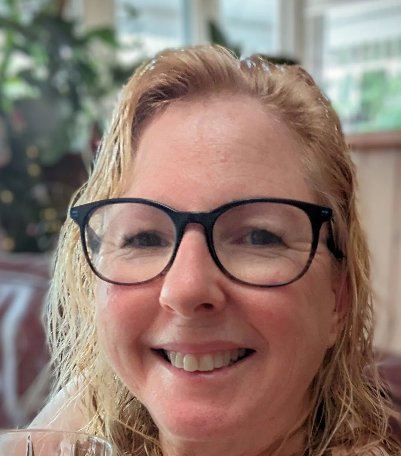 A smiling woman with glasses and red hair indoors with trees and windows in the background.