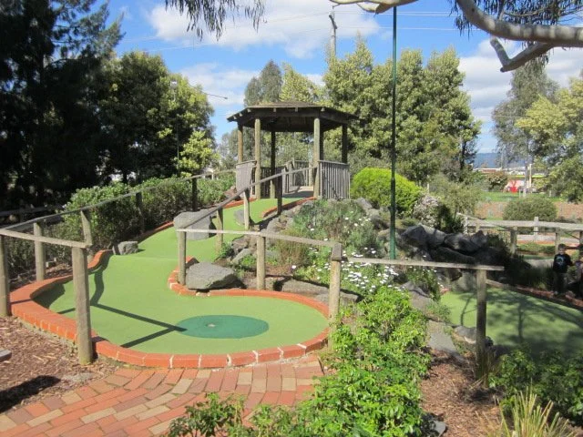 Miniature golf course with a small hill, ramp, and covered seating area surrounded by trees and shrubs.