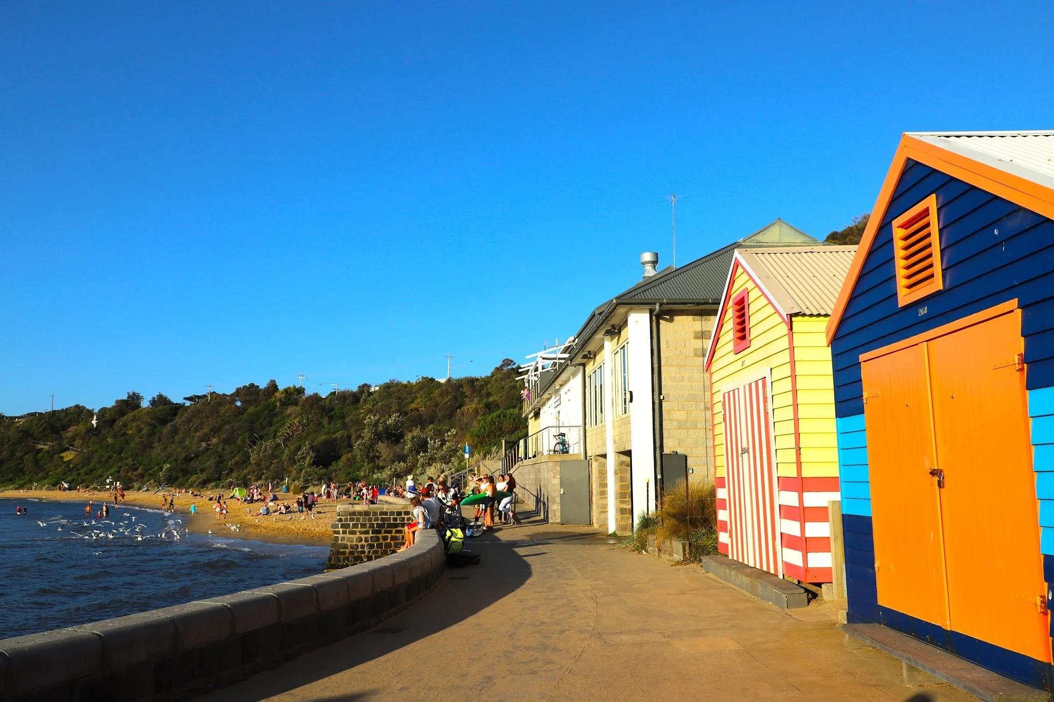 Colorful beach huts along a seaside walkway with people swimming and relaxing on the sandy beach, hillside with greenery in the background, under a clear blue sky.