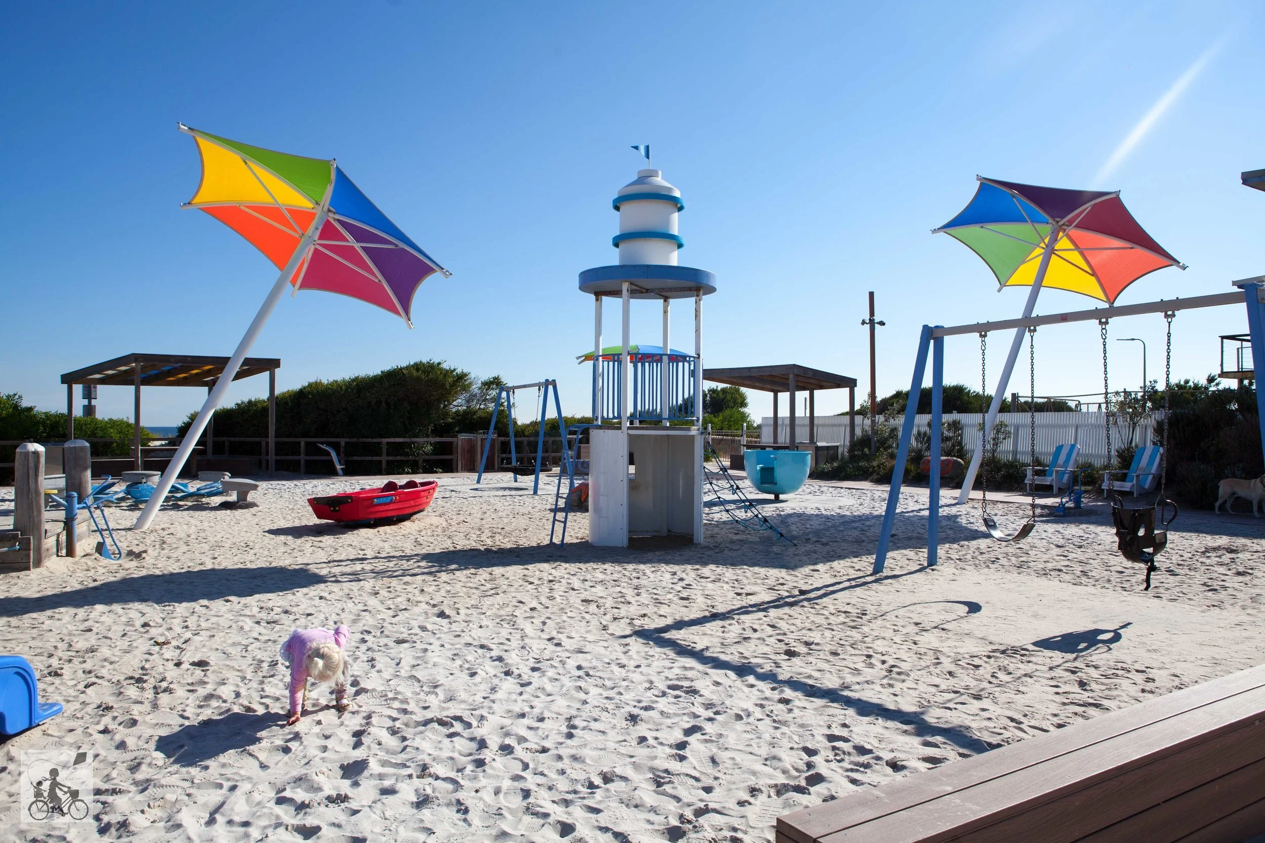 A playground on a sandy beach with colorful umbrellas, swings, a slide, and a small child playing in the sand on a sunny day.
