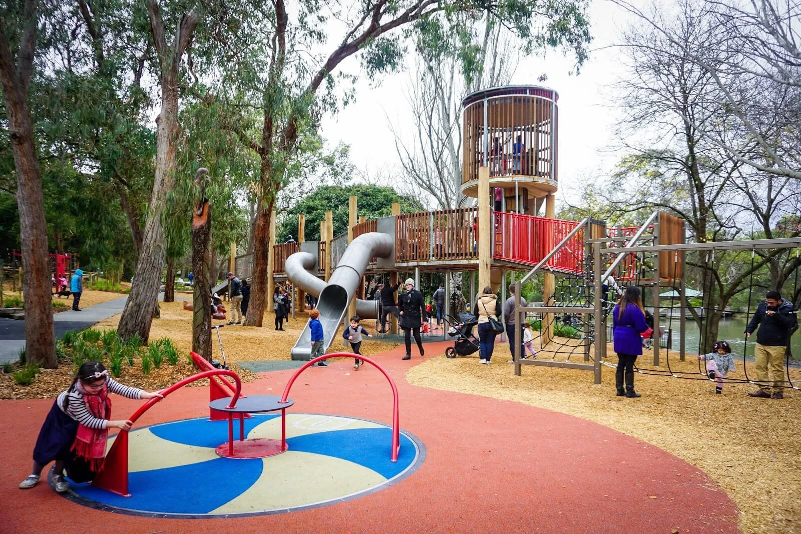 Children and adults playing and walking around a colorful playground with slides, climbing structures, and a tall wooden tower in a park with trees.