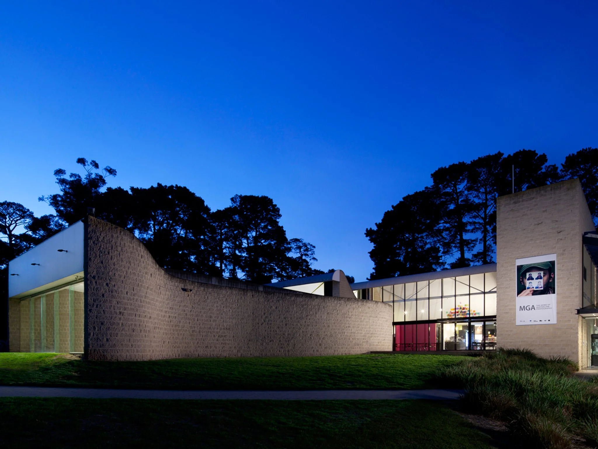 Modern museum building with large glass windows and a poster of an artist on the exterior, surrounded by trees at dusk.