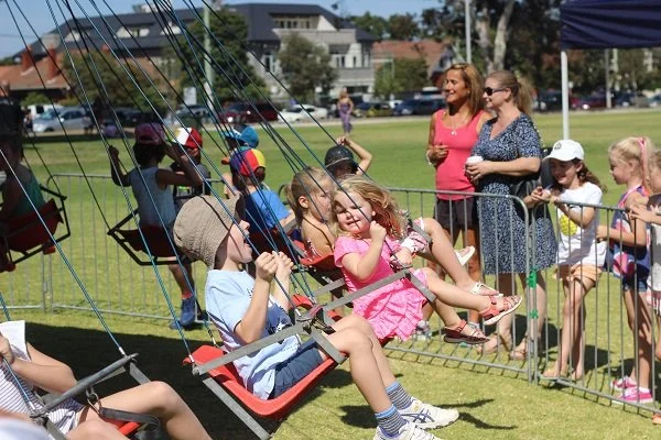 Children on swings at a fair or park, with two women standing nearby watching and smiling.