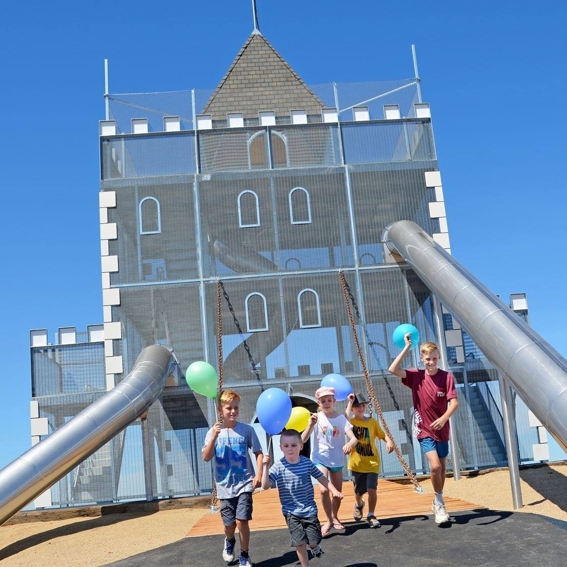 Children playing on a playground in front of a castle-shaped building with slides and a metal slide, holding colorful balloons on a sunny day.