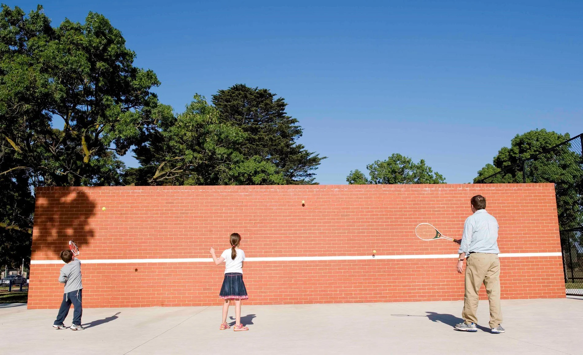 A man and two children playing tennis on a court with a red brick wall, green trees, and clear blue sky in the background.