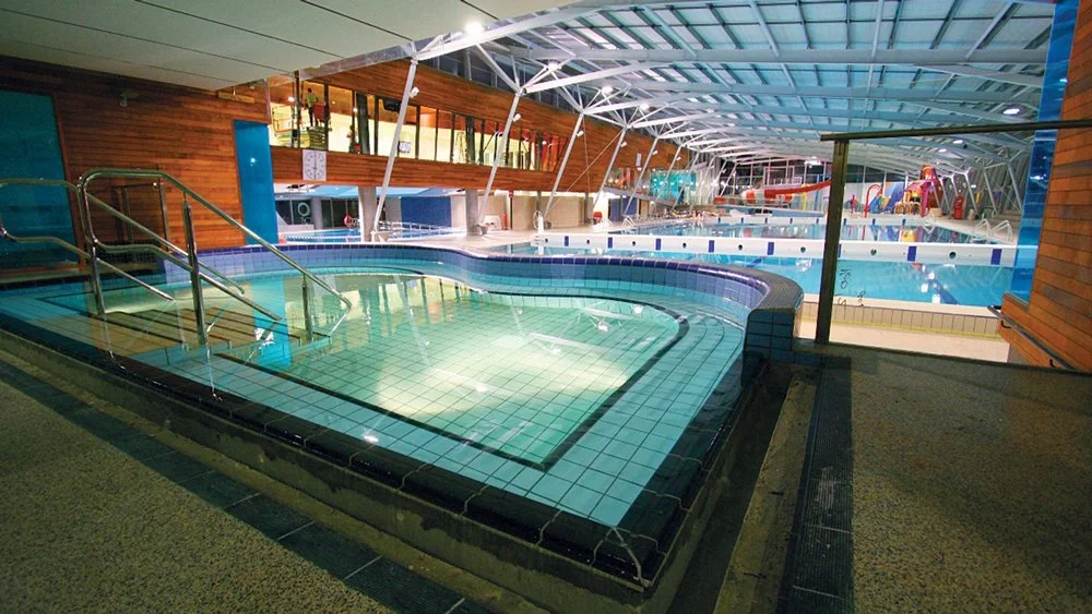 Indoor water park with a hot tub in the foreground and a larger swimming pool area with slides and water features in the background, under a roof structure with glass panels.